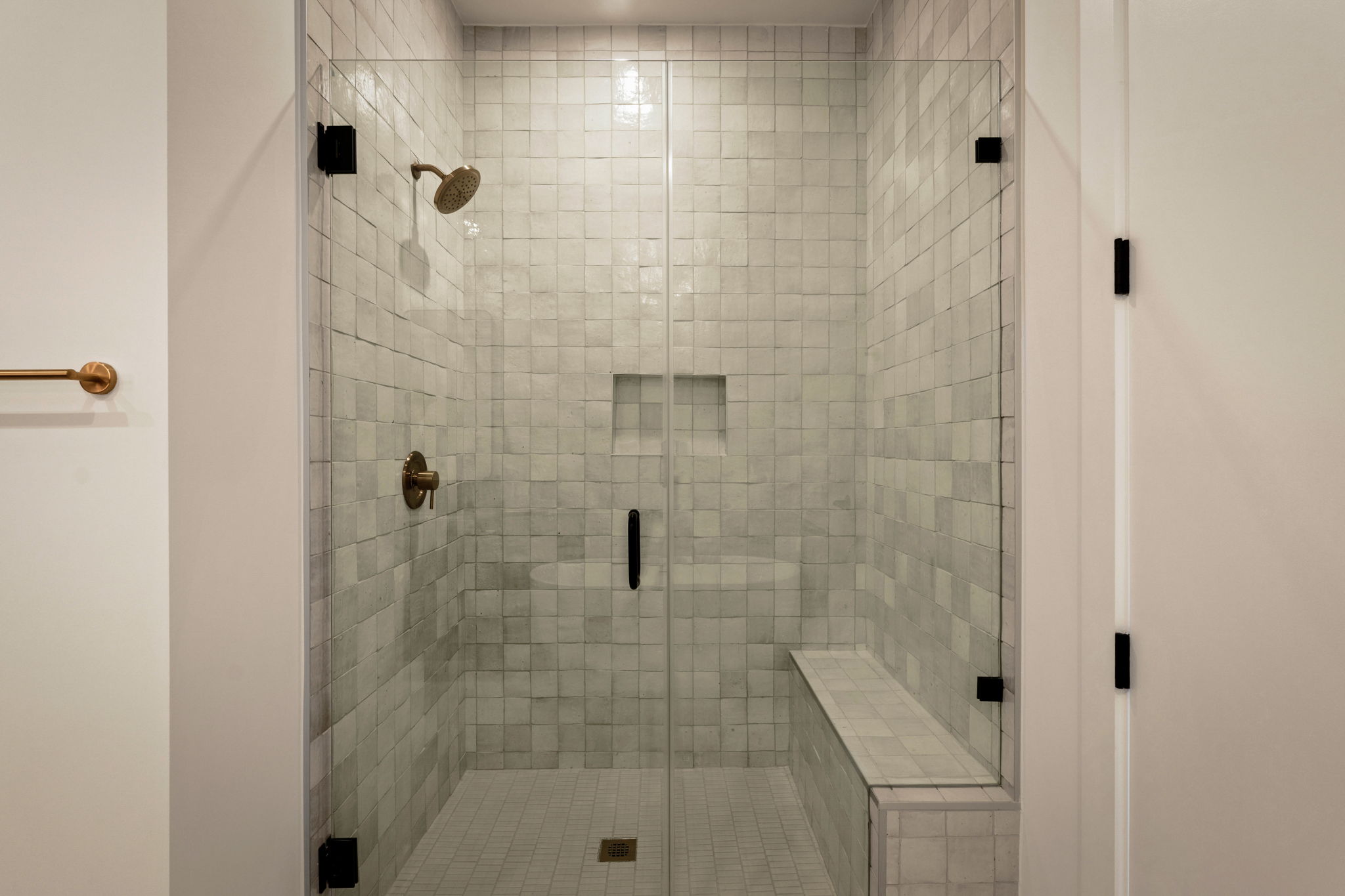 A modern walk-in shower in the Paseo Penthouse features white square tiles, a glass door, built-in bench, recessed shelf, and bronze showerhead and fixtures.