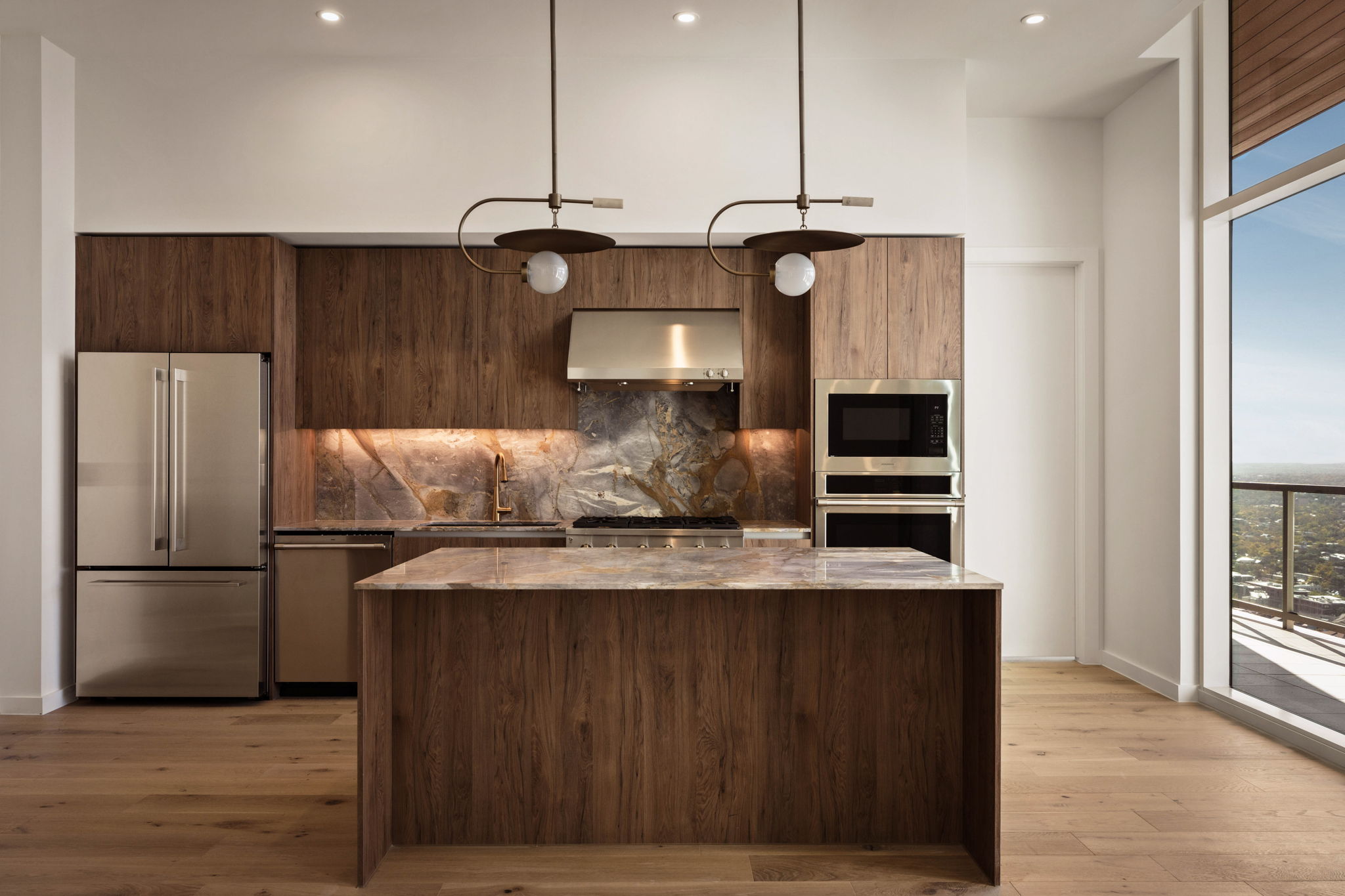 Modern kitchen with wood cabinets, marble countertops, stainless steel appliances, built-in oven and microwave, pendant lights above the island, and large windows offering a city view—perfect for a Paseo Penthouse lifestyle.