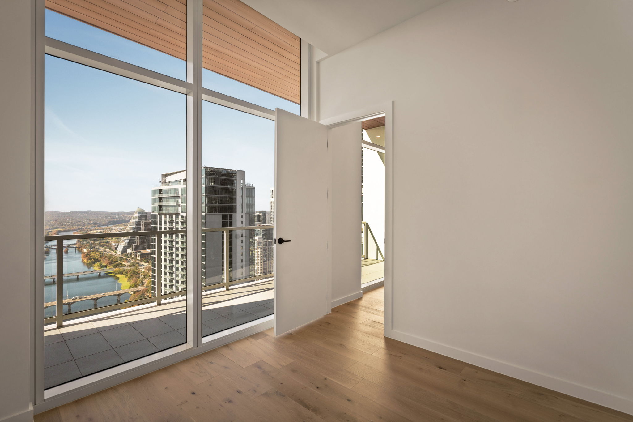 A bright, empty paseo penthouse room with light wood floors, white walls, and floor-to-ceiling windows opening to a balcony overlooking modern high-rise buildings and a river below.