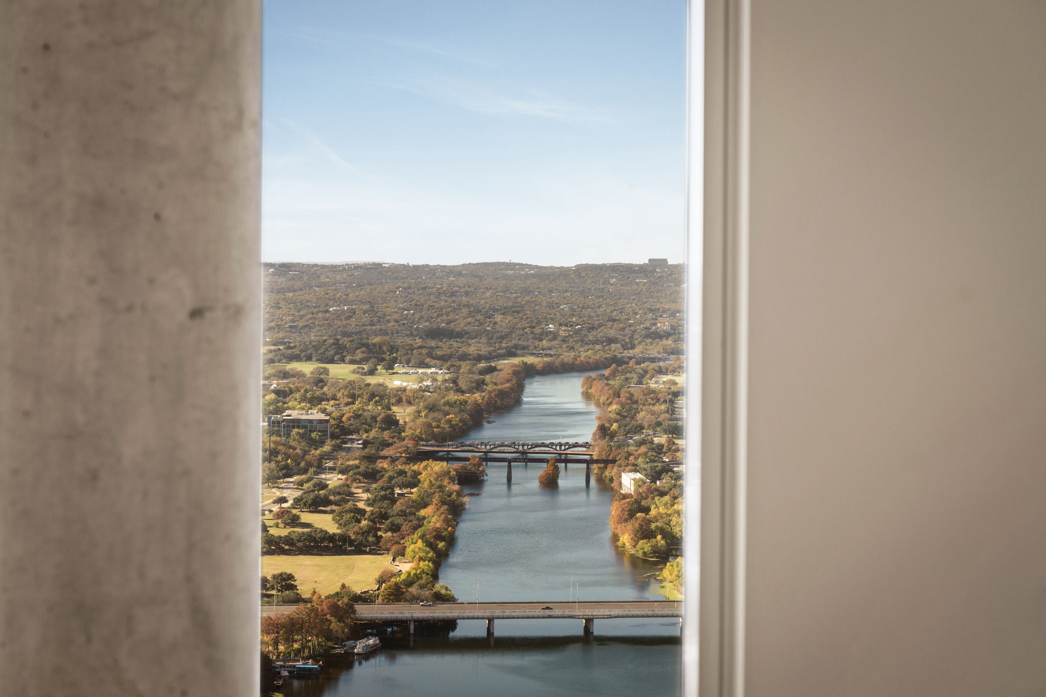View through a window frame in the Paseo Penthouse, showing a river with two bridges, surrounded by trees and hills under a clear blue sky.