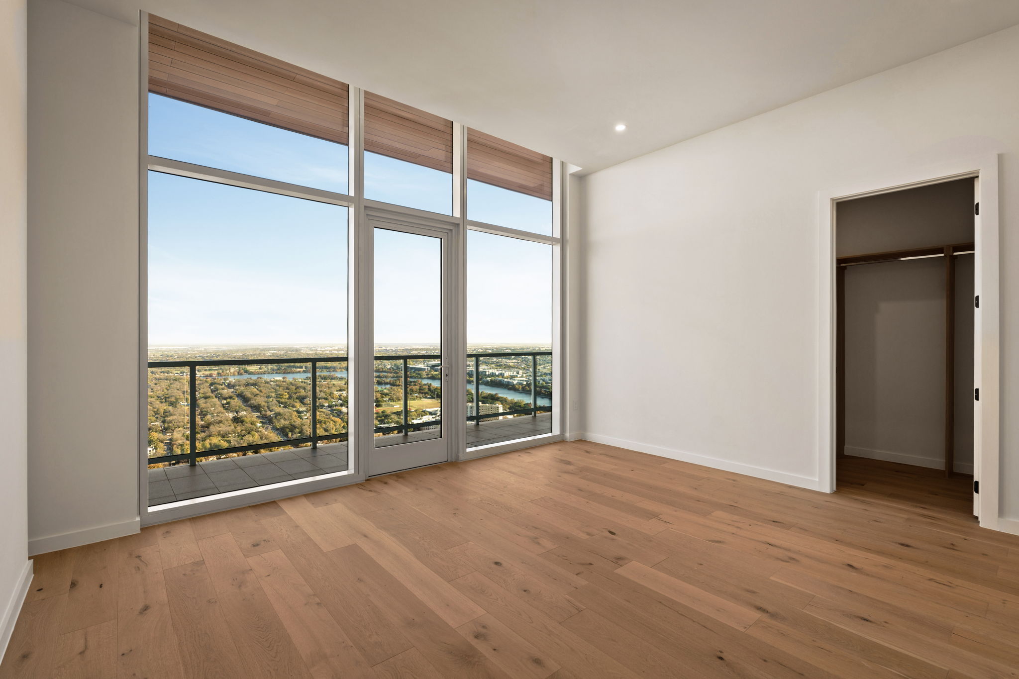 A spacious, empty paseo penthouse room with light wood flooring, large floor-to-ceiling windows, a glass door to a balcony, and an open closet; the windows reveal a scenic view of trees and water.