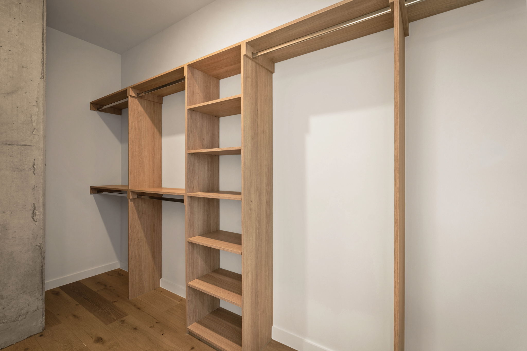 A modern, empty walk-in closet in the Paseo Penthouse features light wood shelves, open hanging rods, and cubby compartments set against white walls and a hardwood floor.