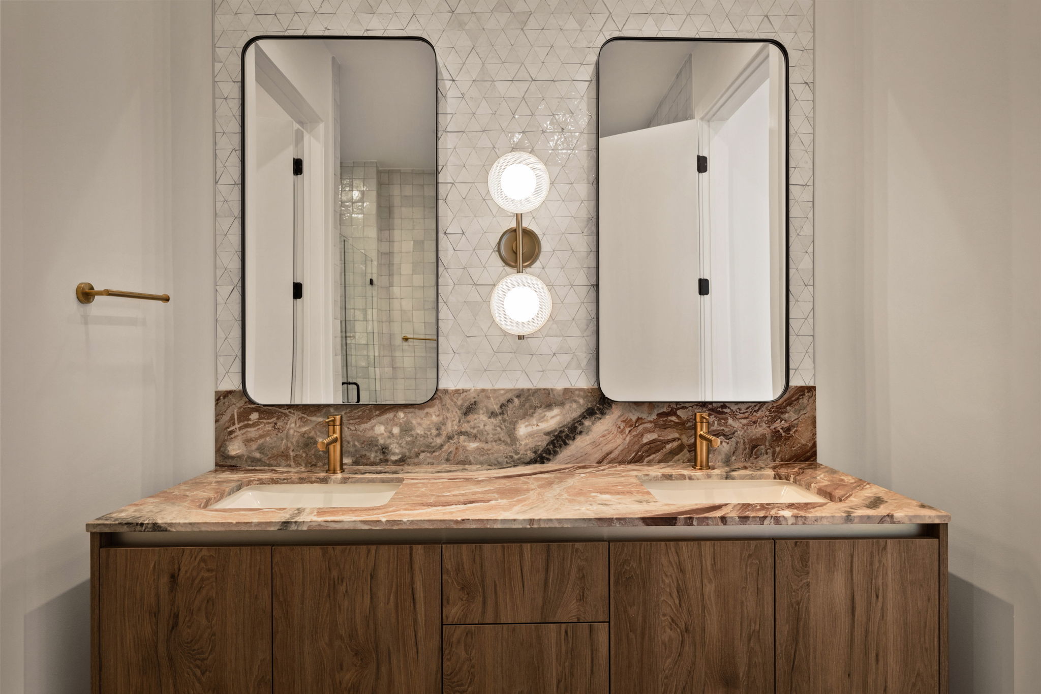 Modern bathroom in the Paseo Penthouse with a double sink vanity, marble countertop, two rectangular mirrors, geometric tile backsplash, and gold fixtures. A central light fixture with two round bulbs is mounted between the mirrors.