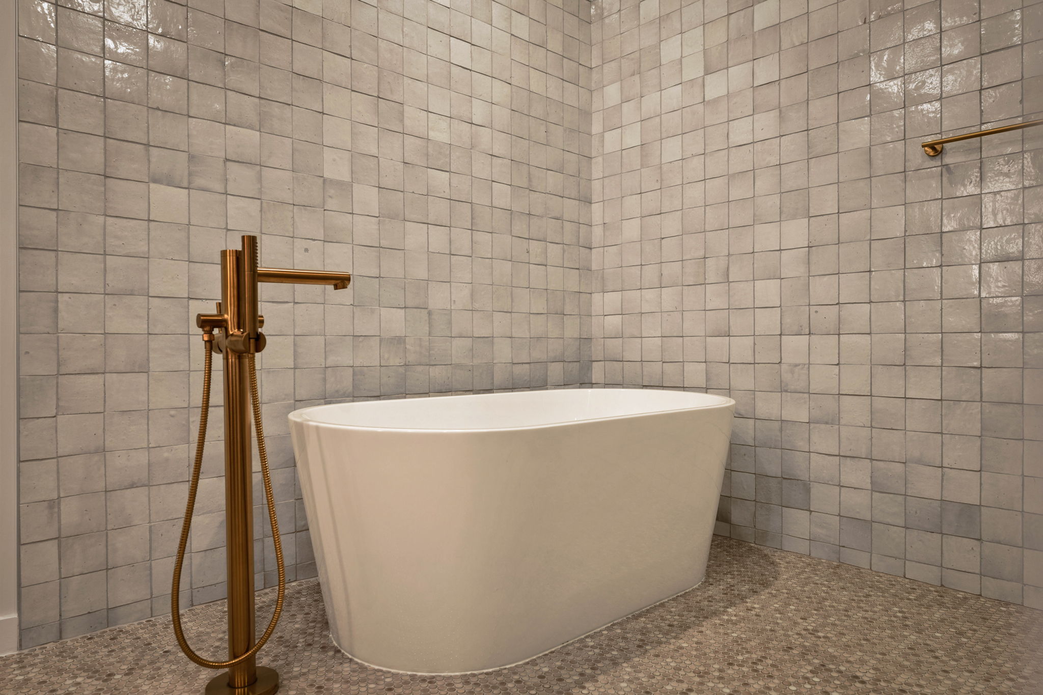 A modern bathroom in the Paseo Penthouse features square light gray tiles, a white freestanding bathtub, and a floor-mounted gold faucet set on a small mosaic tile floor.