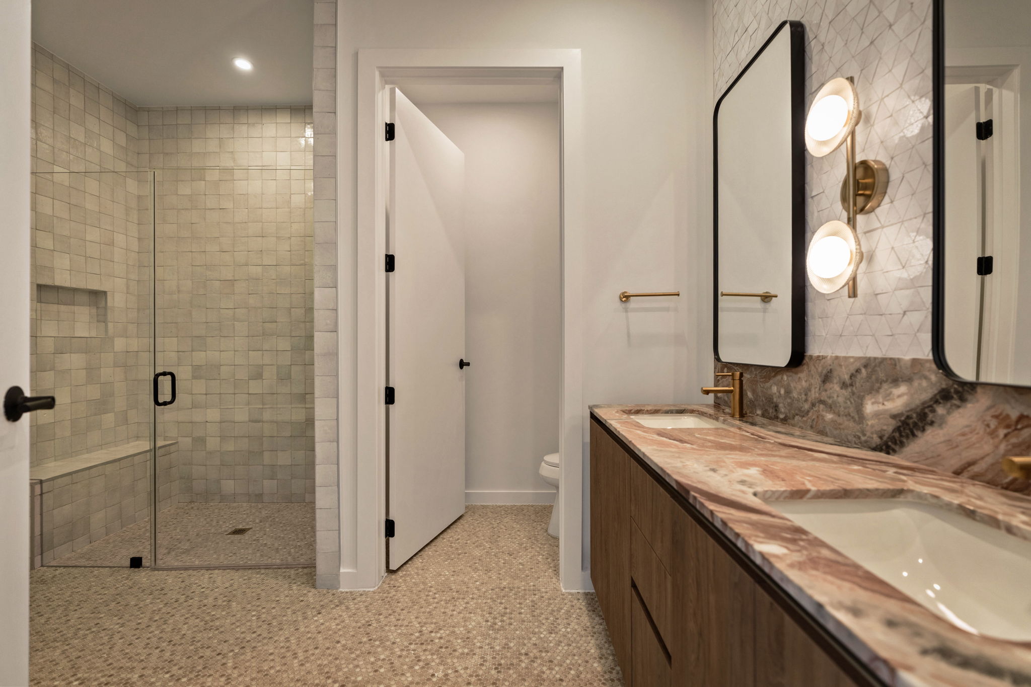 Modern bathroom in the Paseo Penthouse with a double sink vanity, marble countertop, two rectangular mirrors, gold fixtures, walk-in shower with glass door, and neutral tiled floor and walls. A door to a toilet area is slightly open.