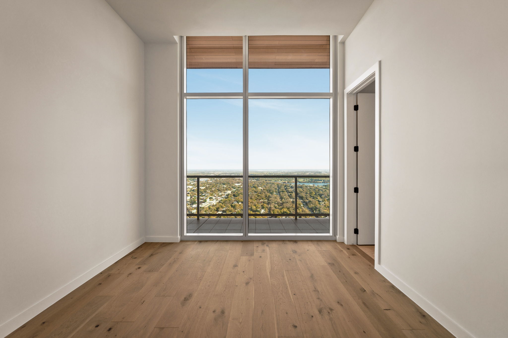 A bright, empty Paseo Penthouse room with light wood floors, white walls, and large floor-to-ceiling windows opening to a balcony with a scenic city and sky view. A doorway is visible on the right.