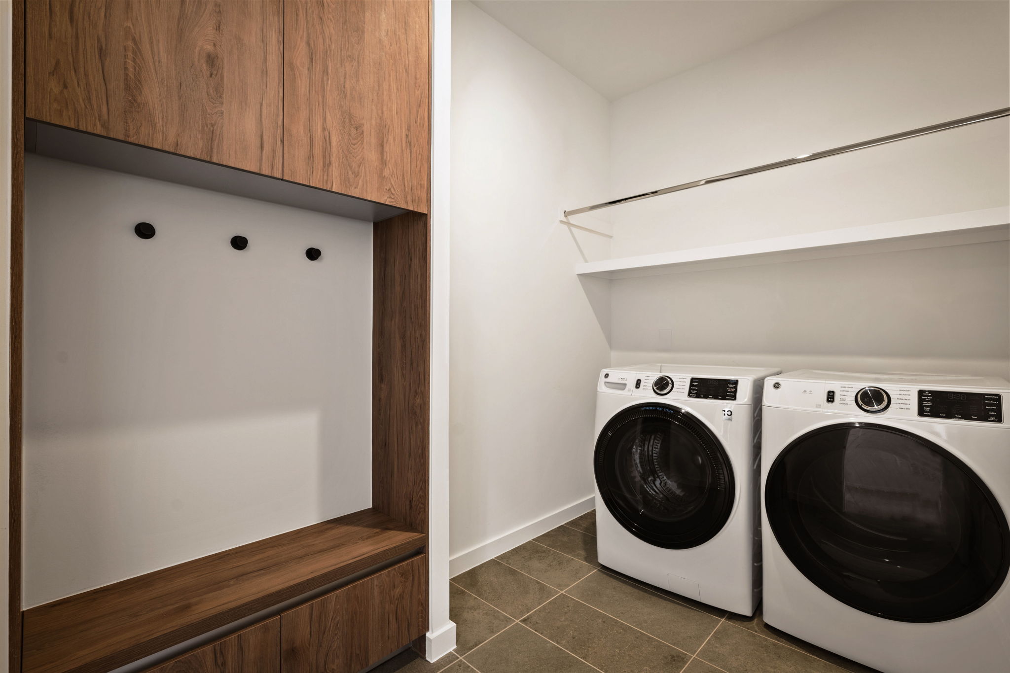 Modern laundry room in the Paseo Penthouse with a front-loading washer and dryer, built-in wooden bench with coat hooks, white walls, overhead shelving, and tiled floor. The space looks clean and minimalistic.