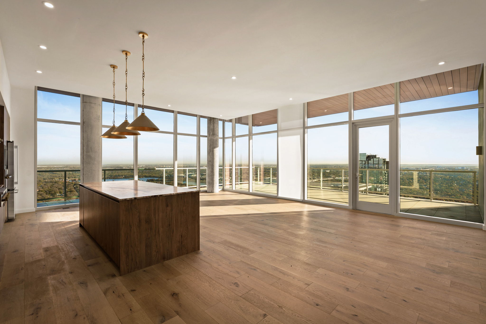 Spacious, modern paseo penthouse with large floor-to-ceiling windows, wooden floors, a central island with pendant lights, and a balcony offering panoramic views of the landscape and distant buildings.