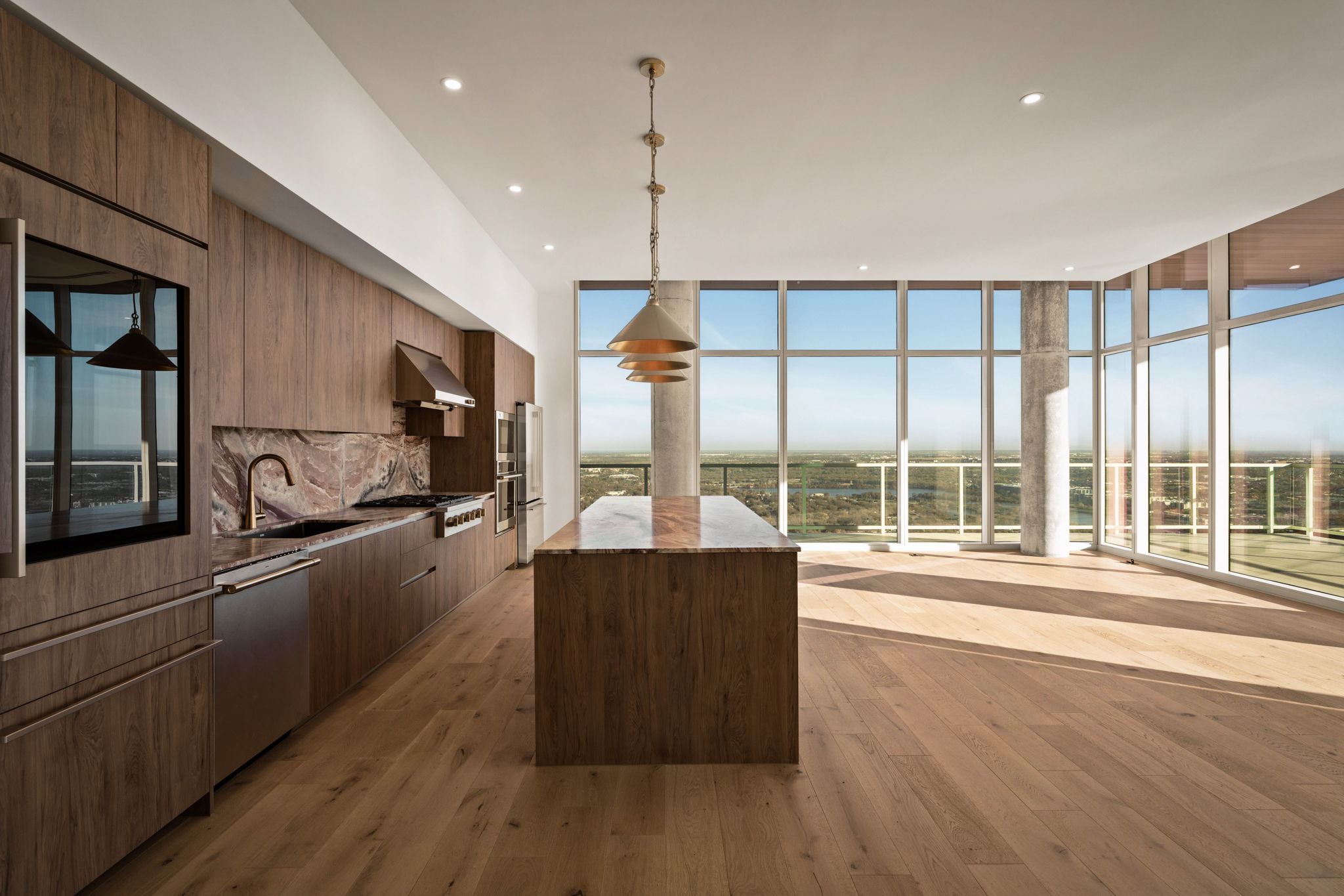 Spacious modern kitchen in the Paseo Penthouse with wood cabinets, marble backsplash, large island, built-in appliances, pendant lights, and floor-to-ceiling windows offering a panoramic view and abundant natural light.