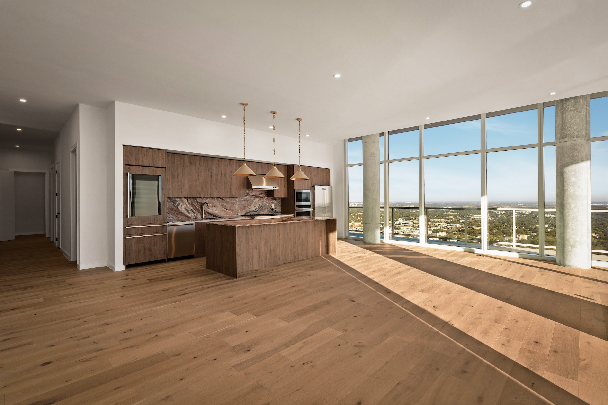 A modern, spacious Paseo Penthouse apartment with light wood floors, an open kitchen featuring brown cabinets and island, large floor-to-ceiling windows, and a panoramic view of the city outside.