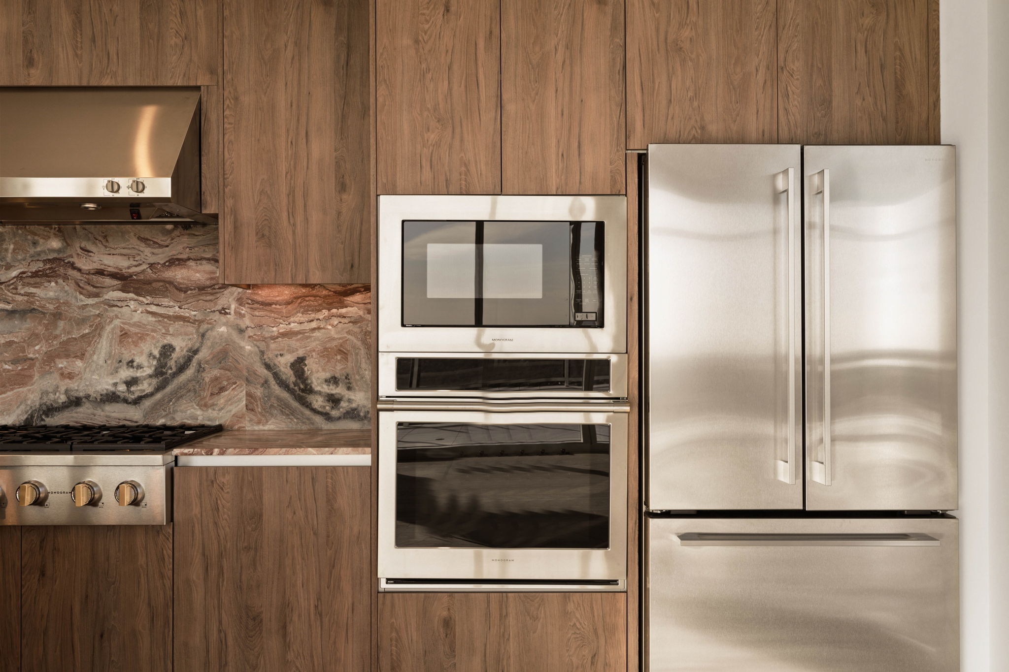 Modern kitchen in the Paseo Penthouse with wood cabinetry, marble backsplash, stainless steel appliances including a double-door fridge, built-in microwave, oven, and gas stove with vent hood. Clean, contemporary design.