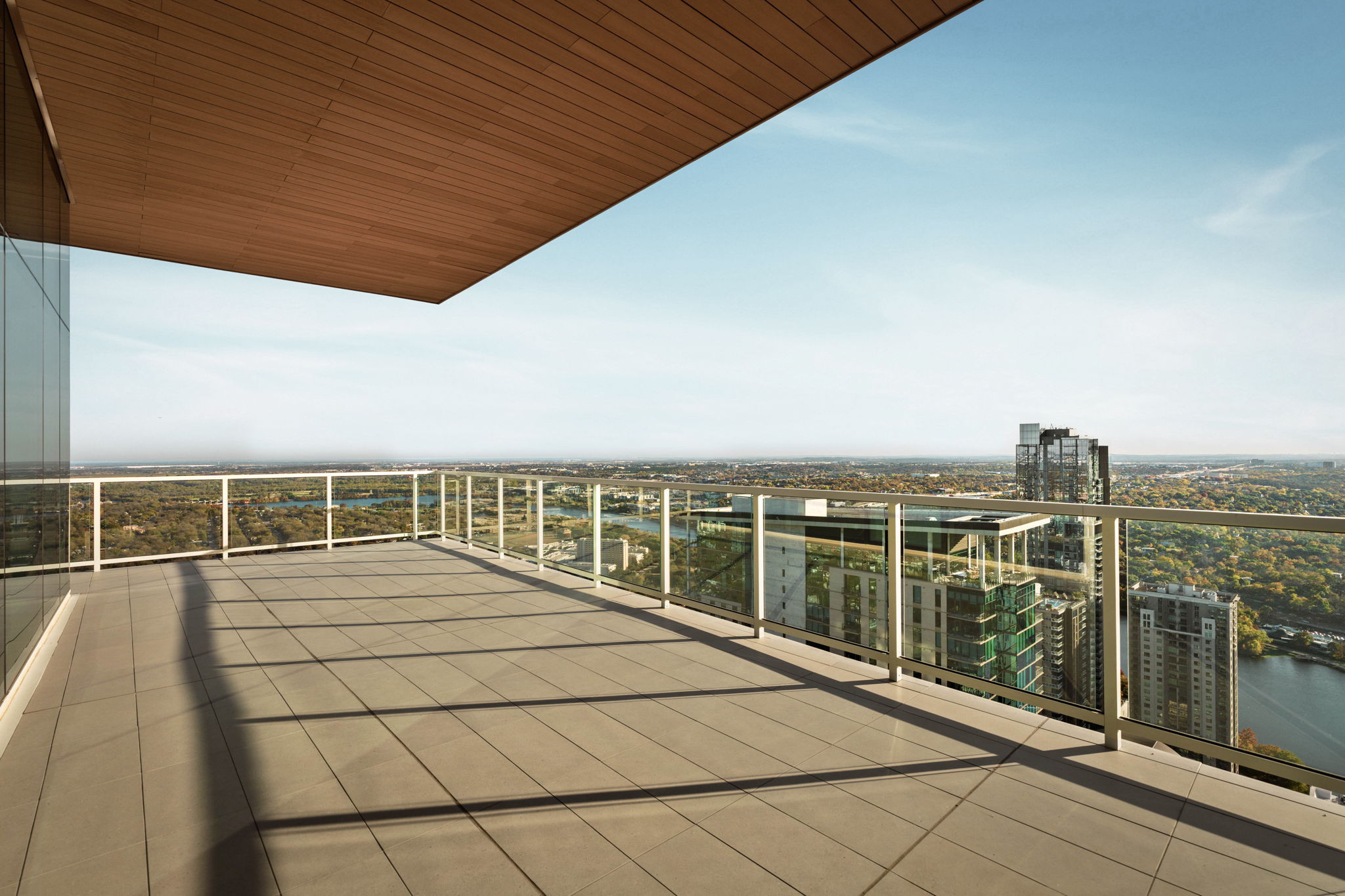 A spacious, modern balcony with glass railings and a tiled floor offers a panoramic view of the city skyline and distant greenery—an ideal retreat in the luxurious Paseo Penthouse under a clear blue sky.