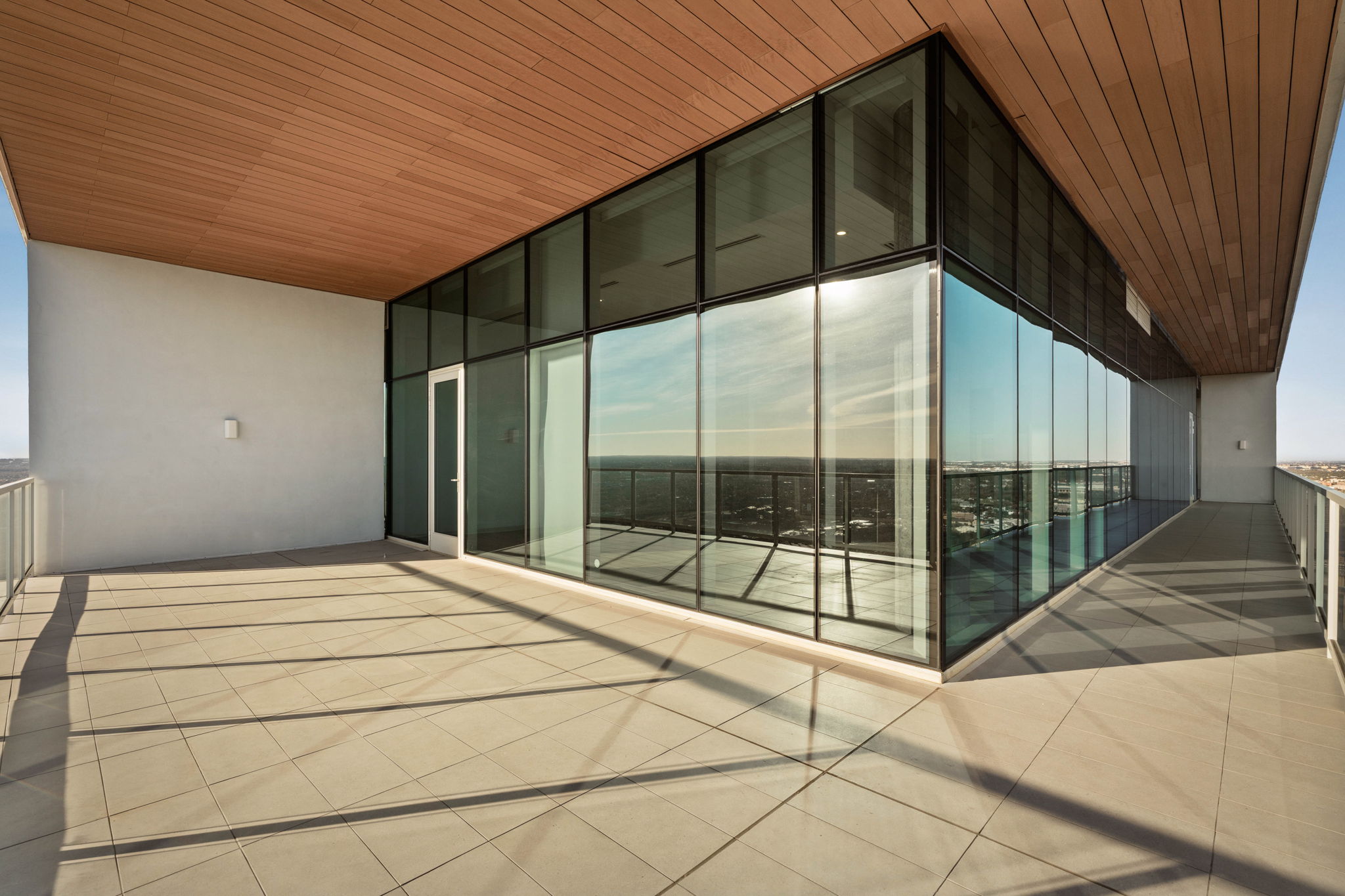A modern, empty Paseo Penthouse balcony with large glass windows and doors, a tiled floor, and a wooden ceiling, offering expansive city views under clear skies.