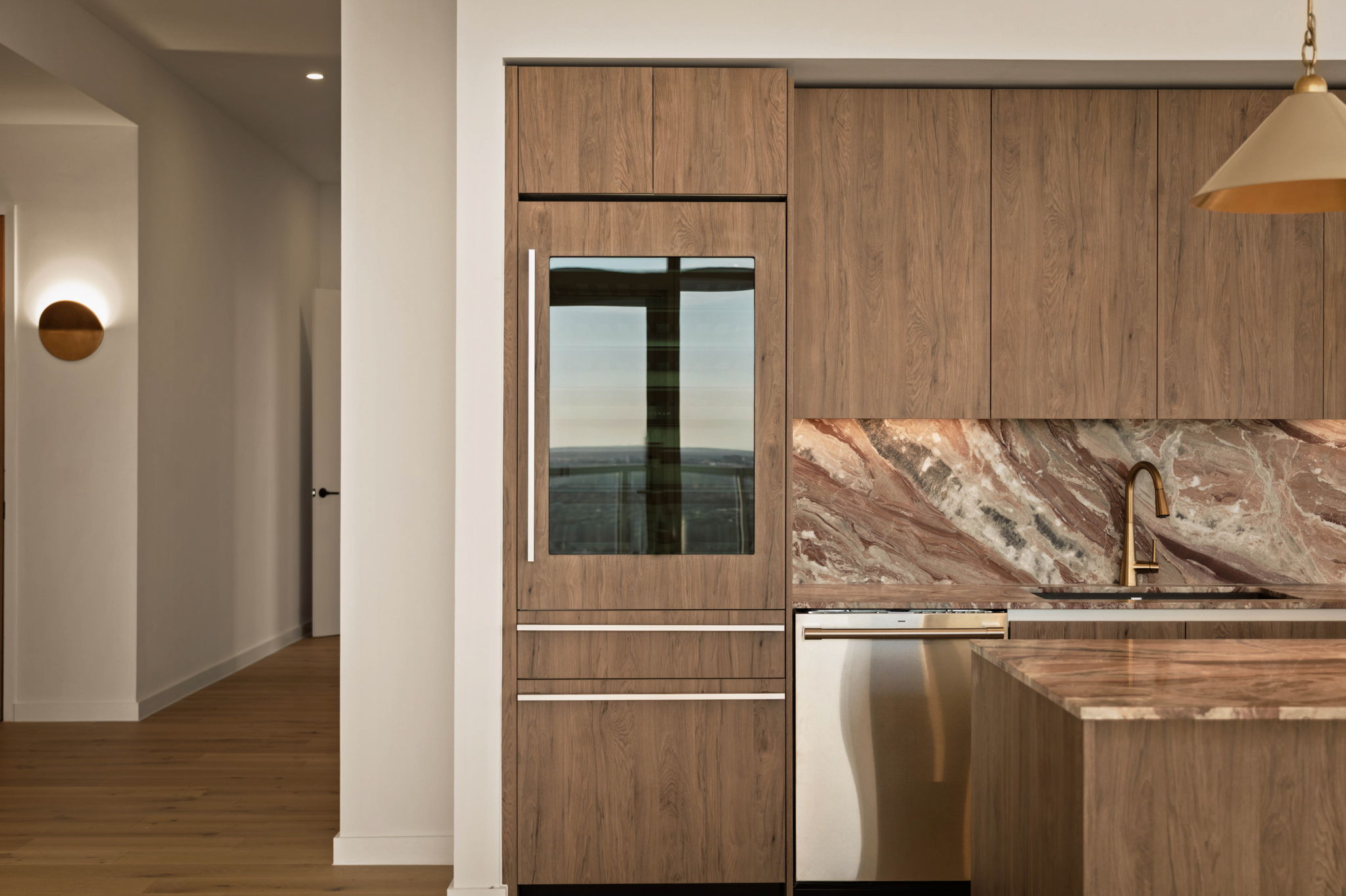 Modern kitchen in the Paseo Penthouse features wood-paneled cabinets, a built-in refrigerator, marble backsplash and countertop, gold faucet, stainless steel dishwasher, and a pendant light. A hallway with wood flooring is visible on the left.