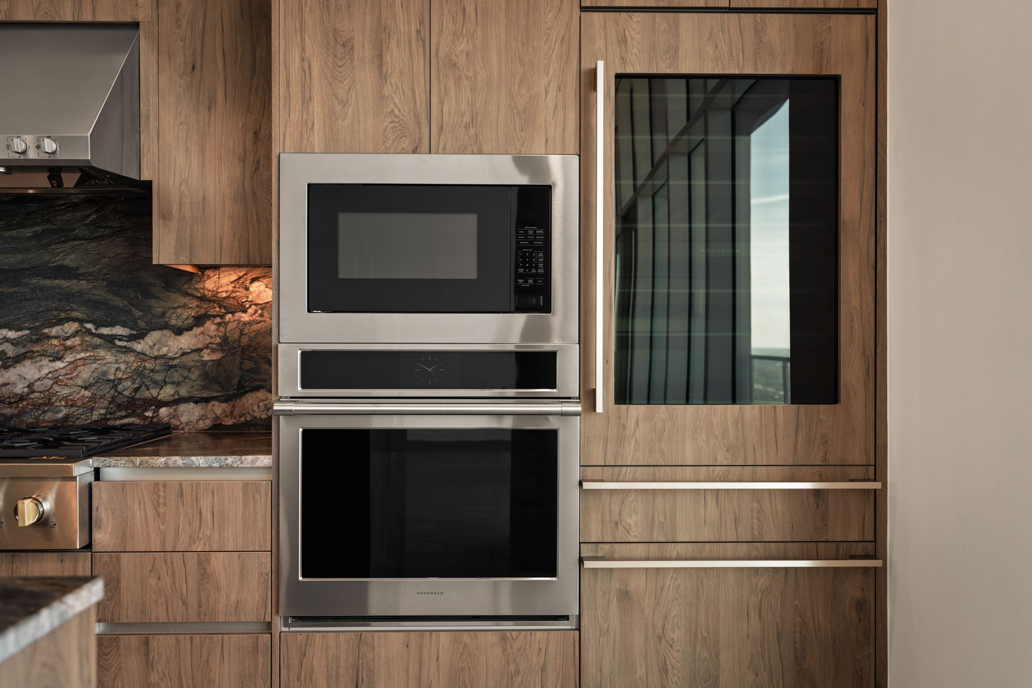 Modern kitchen with wooden cabinets, built-in stainless steel microwave and oven, and a glass-front refrigerator. The marble backsplash with unique veining adds elegance to this stunning Paseo Penthouse space.
