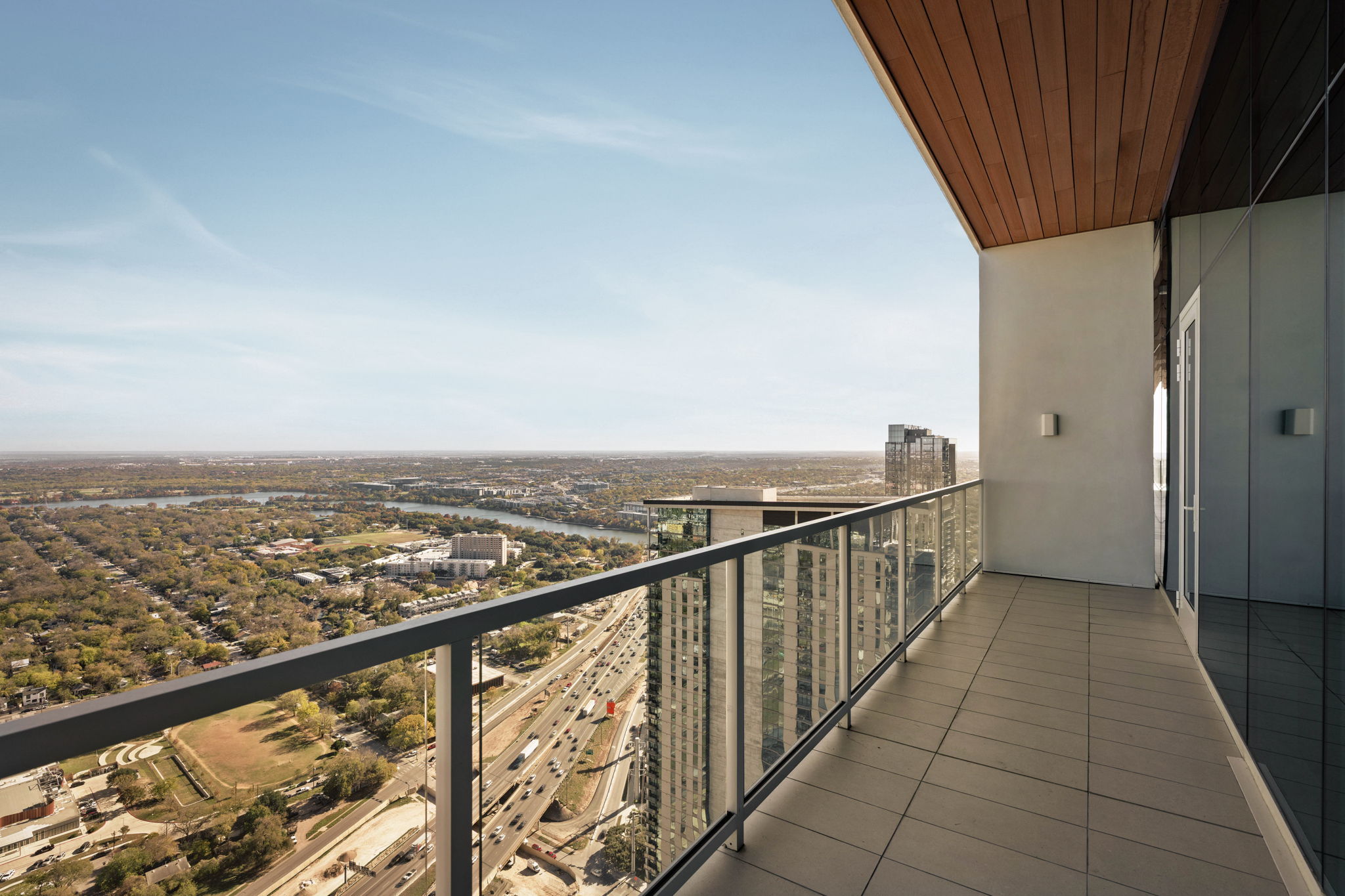 A modern high-rise paseo penthouse balcony with glass railing overlooks a cityscape, featuring a wide view of buildings, roads, greenery, and a river under a clear blue sky.