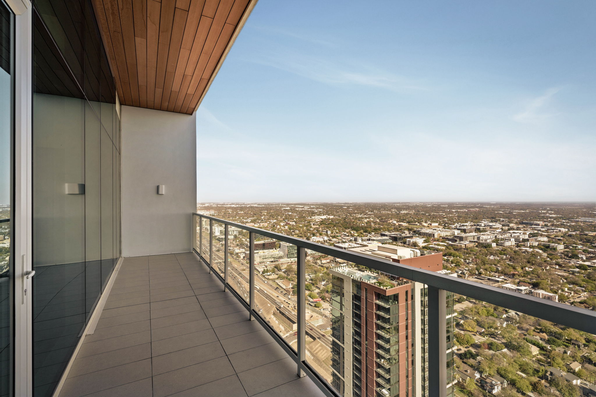 A high-rise paseo penthouse balcony with glass railing and tiled floor overlooks a cityscape with buildings, roads, and greenery under a clear blue sky.