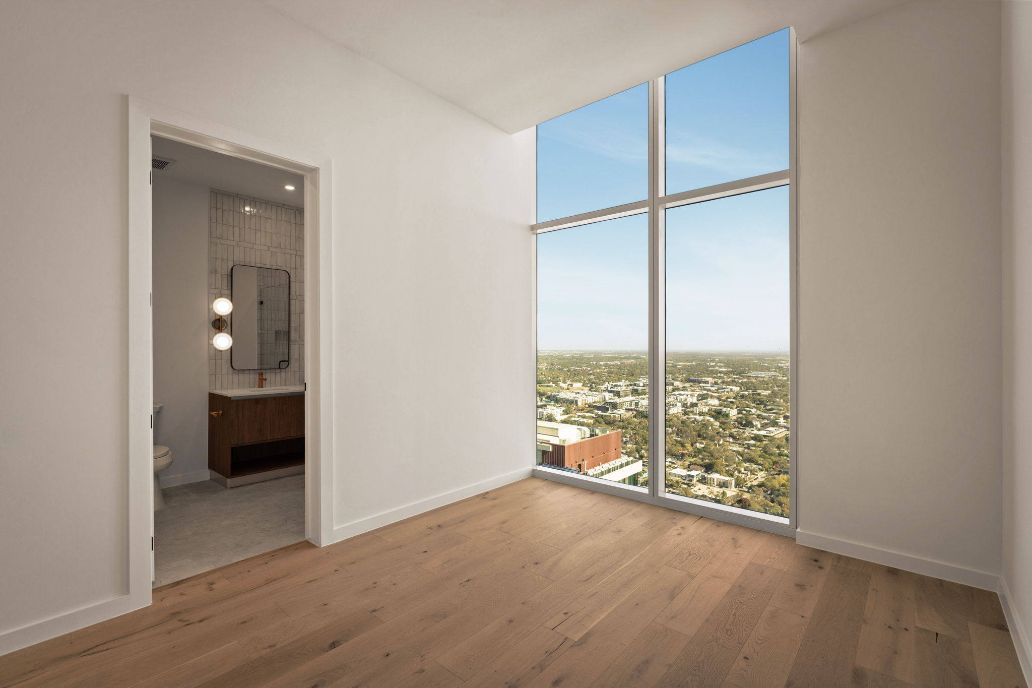 A bright, empty room in the Paseo Penthouse features light wood floors, a large floor-to-ceiling window with stunning city views, and an open doorway leading to a modern bathroom with a lit mirror and tiled walls.