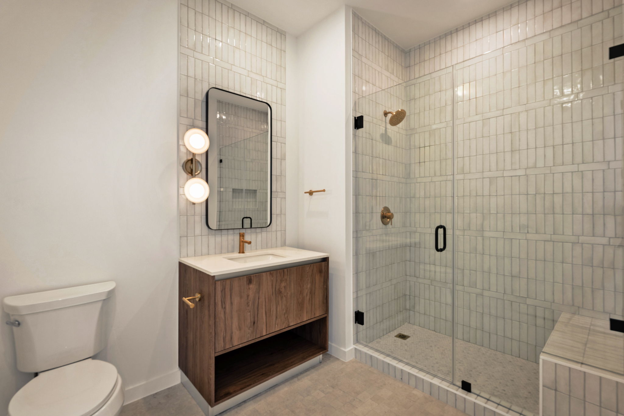 Modern bathroom in the Paseo Penthouse with a glass-enclosed shower, white subway tile walls, wooden vanity with gold faucet, rectangular mirror, wall-mounted lights, and a white toilet. The design is clean and minimal.