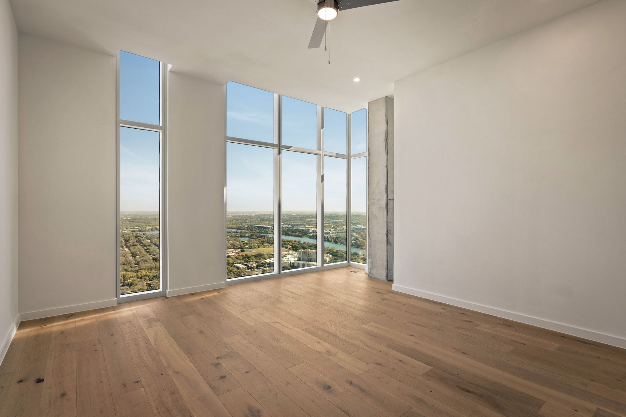 Bright, empty Paseo Penthouse room with light wood flooring, tall floor-to-ceiling windows offering a panoramic city view, white walls, a ceiling fan, and a visible concrete column in the corner.