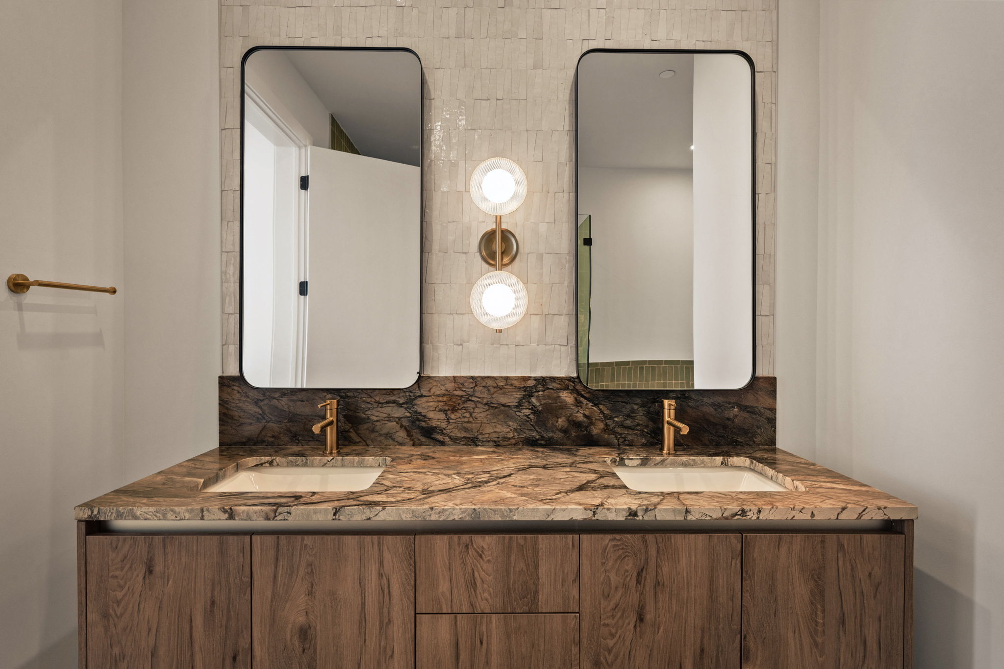 Modern bathroom in the Paseo Penthouse with a double sink vanity, marble countertop, wooden cabinets, two rectangular mirrors, gold faucets, and a wall-mounted light fixture between the mirrors. White tiled wall backdrop.