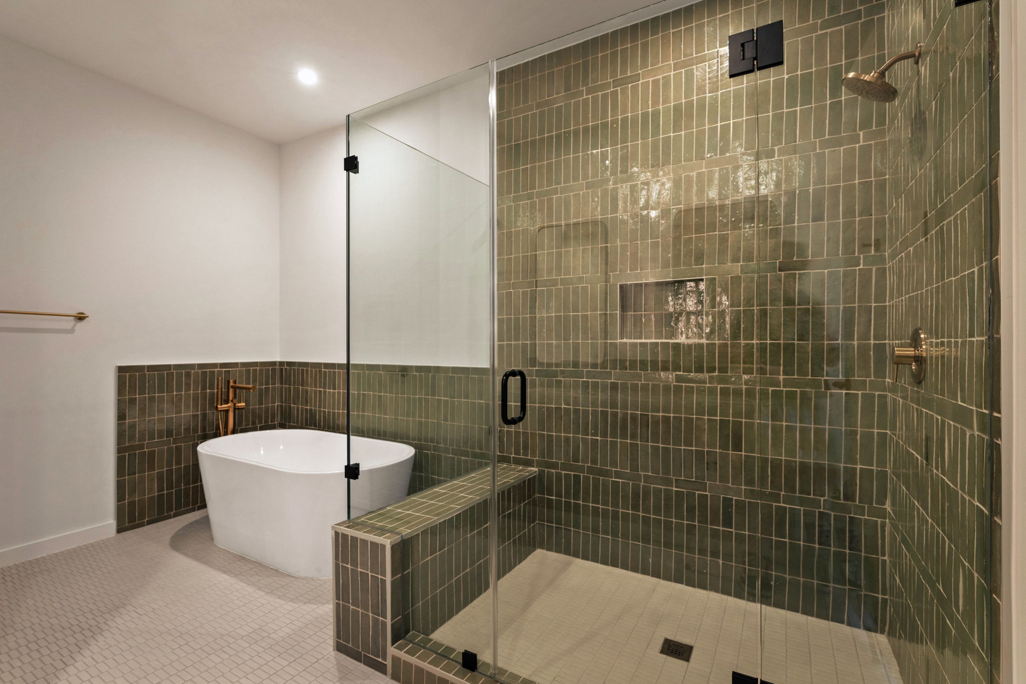 Modern bathroom in the Paseo Penthouse with a glass-enclosed shower featuring green tiles, a built-in bench, and recessed shelf. A white freestanding bathtub and gold fixtures complement the light-colored walls and floor tiles.