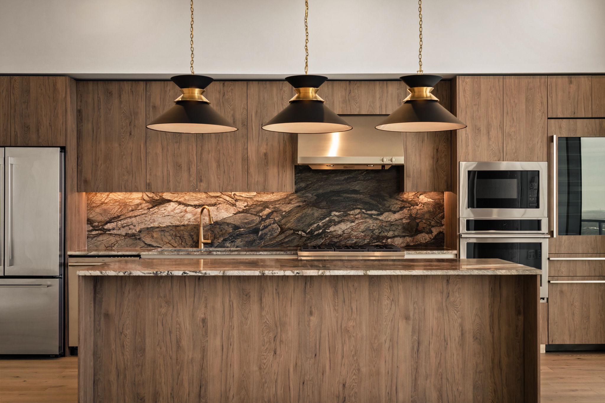 Modern kitchen in the Paseo Penthouse with wood cabinets, a marble island, three black and gold pendant lights, stainless steel appliances, and a dramatic stone backsplash featuring swirling patterns behind the stove and sink.