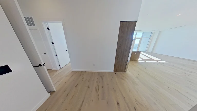 Minimalist, unfurnished room with light wood floors, white walls, and a partially open wooden sliding door. Sunlight streams through large windows in an adjacent, bright living area. Two closed doors are visible to the left.