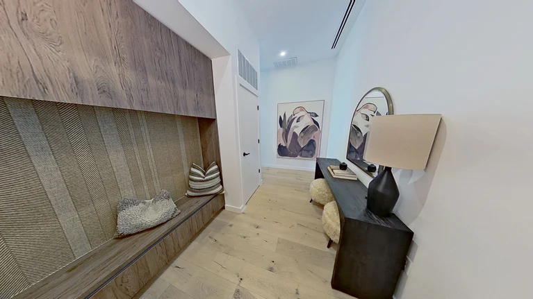 A modern hallway with light wood flooring, built-in bench with pillows, dark console table, round mirror, lamp, decorative objects, and abstract art on the far wall.