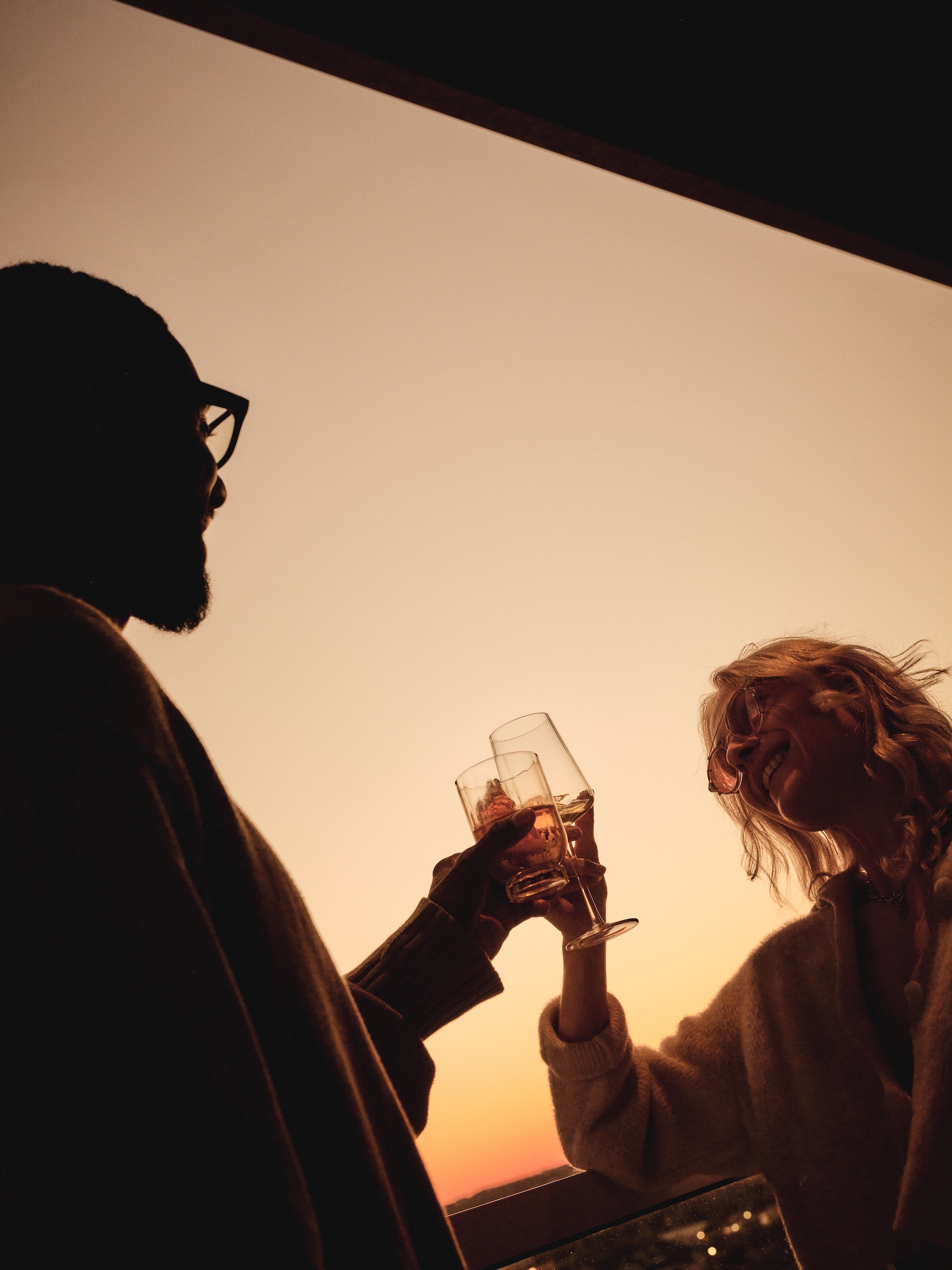 Two people clinking glasses and smiling at sunset, silhouetted against a warm, orange sky at the downtown Austin Paseo apartment pool —capturing a moment of shared wellness and joy.