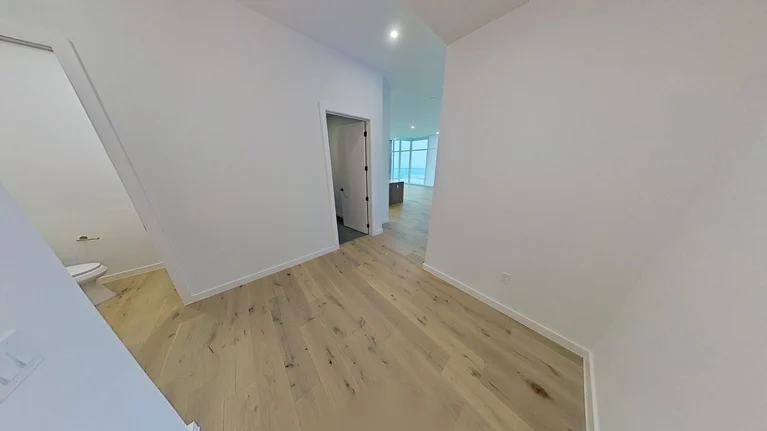 A hallway with light wood flooring and white walls, leading to rooms with open doorways. Recessed ceiling lights are visible, and natural light comes from windows in the distant room. A toilet is partially visible on the left.