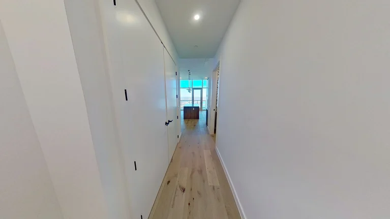 A narrow hallway with light wooden floors, white walls, and closed white doors on the left, leading to a room with large windows and visible outdoor light.